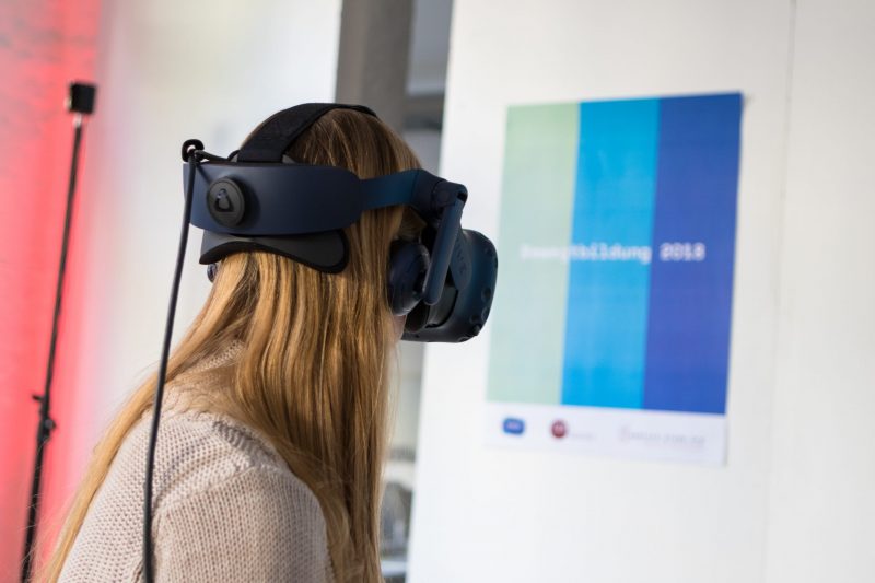 Frau mit VR-Brille, im Hintergrund ein Poster mit den Bewegtbildungsfarben mint, blau und dunkelblau und dem Schriftzug "Bewegtbildung 2018"