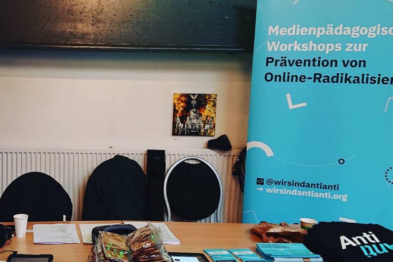Auf dem Bild erkennt man den AntiAnti-Stand auf dem Berliner Jugendforum 2019. Es ist ein Tisch zu sehen, auf dem Sticker, iPads und Beutel ausliegen. Im Hintergrund steht der AntiAnti-Aufsteller.