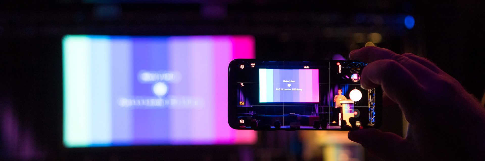 Kamerabildschirm filmt eine große Leinwand ab, auf dem das Logo und die Farben von Bewegtbildung.net zu sehen sind.