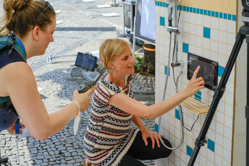 Frau erklärt das Exponat der Singdusche, andere Frau filmt mit dem Handy