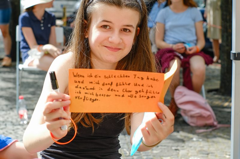 Mädchen hält beschriebene Moderationskarte in die Kamera, auf der sie schreibt, dass ihr Musik hilft, wenn sie schlechte Laune hat.